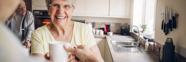 Senior woman wearing glasses standing in her kitchen. She is holding a cup and smiling. Relaxed, happy as she looks at someone just seen in the left of the frame.