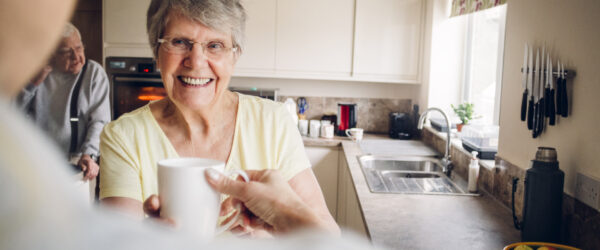Senior woman wearing glasses standing in her kitchen. She is holding a cup and smiling. Relaxed, happy as she looks at someone just seen in the left of the frame.